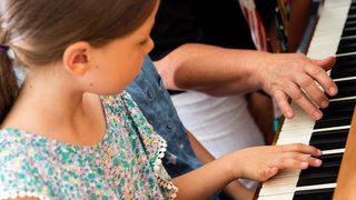 Mädchen im Grundschulalter am Klavier und die rechte Hand einer erwachsenene Person. Das Mädchen schaut sich die Fingerbewegungen ab.