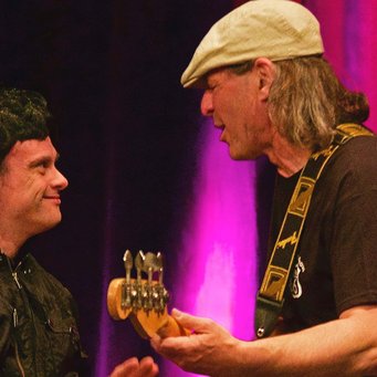 Zwei Männer rocken gemeinsam. Der eine spielt E-Gitarre, der andere trägt eine schwarze Lockenperücke