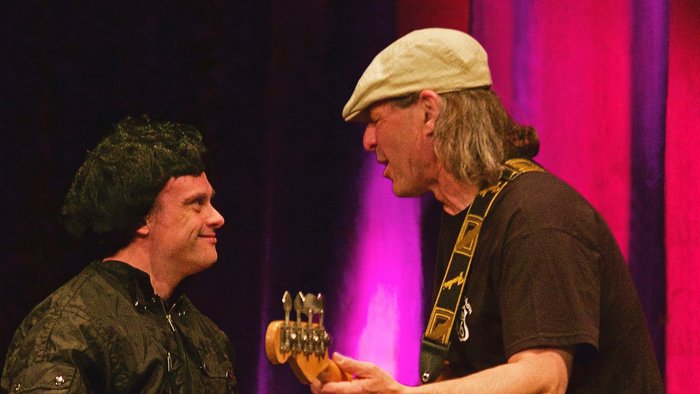 2 Männer rocken gemeinsam auf einer Bühne, der eine trägt eine schwarze Lockenperücke, der andere spielt E-Gitarre