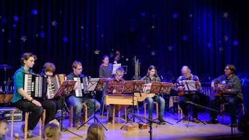 Jugendliche und Erwachsene mit und ohne Behinderung sitzen im Halbkreis auf der Bühne. Sie spielen Akkordeon, Xylofon, Gitarren und Kontrabass