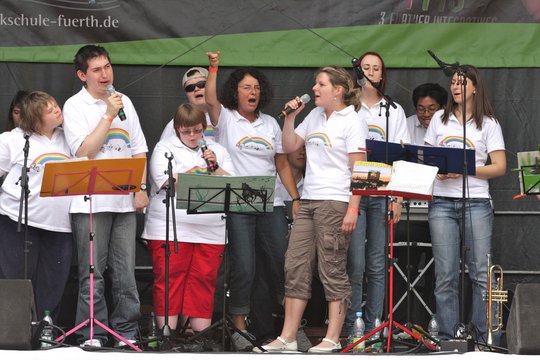 Sänger:innen mit und ohne Behinderung auf der Bühne, sie tragen T-Shirts mit Regenbogen