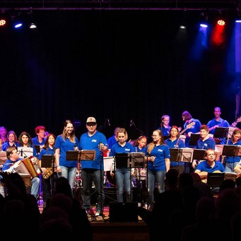 Vollgas Connected: rund 30 Musiker:innen im Festival-T-Shirt stehen und sitzen im Scheinwerferlicht auf der Bühne