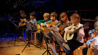 mehrere Kinder mit Gitarren sitzen in einer Reihe auf einer Bühne
