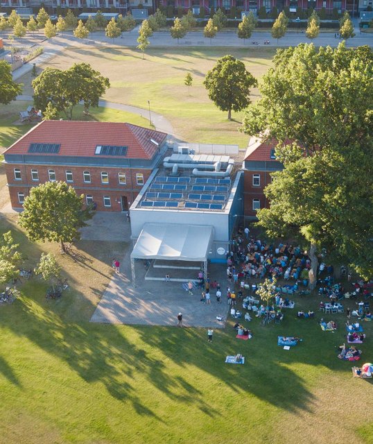 Musikschulgebäude aus der Luft, Bäume werfen lange Schatten, Menschen picknicken bei einem Open-Air-Konzert