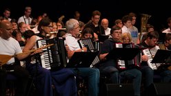 viele Akkordeonist:innen sitzen auf einer Bühne in der ersten Reihe eines großen kunterbunten Orchesters