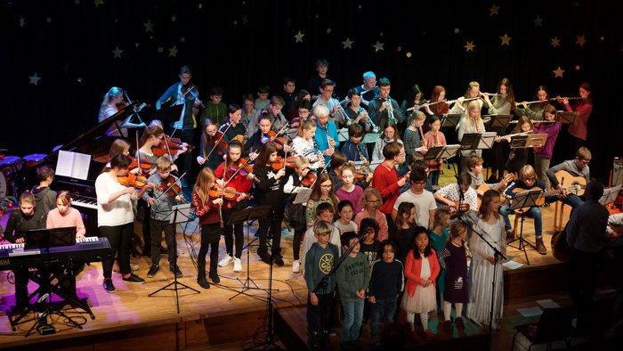 Bühne voller Kinder und Jugendlicher mit verschiedenen Instrumenten, Kamera blickt von oben auf die Bühne