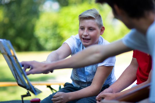 3 Menschen sitzen im Freien vor einem Notenständer, 2 deuten auf die Noten