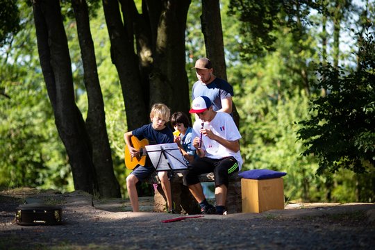 Jugendliche mit Gitarre und Blockflöte auf einer Bank, umringt von Bäumen