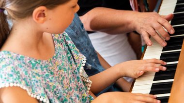 Mädchen sitzt am Klavier, links neben ihr sieht man eine Hand. Das Mädchen schaut sich die Fingerbewegungen ab.